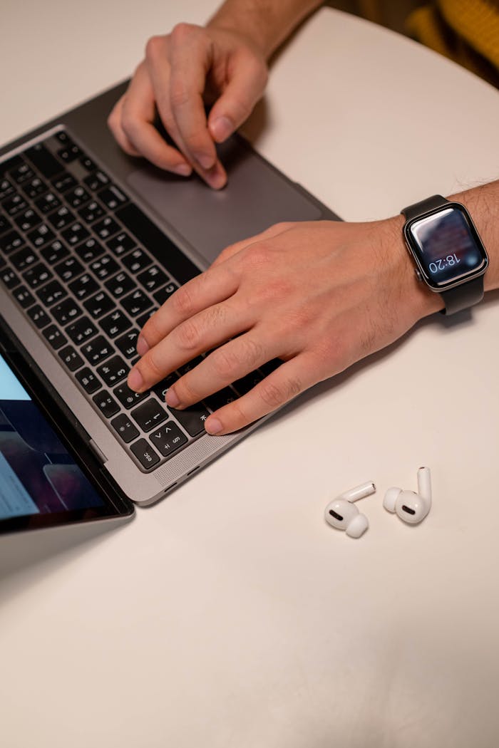 our-story Close-up of a man typing on a laptop while wearing a smartwatch, with earbuds on a table.